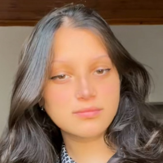 Ana, aluna de direito, posando em um ambiente interno com iluminação suave. Ela tem cabelos escuros e lisos, e usa uma blusa escura com estampa discreta.