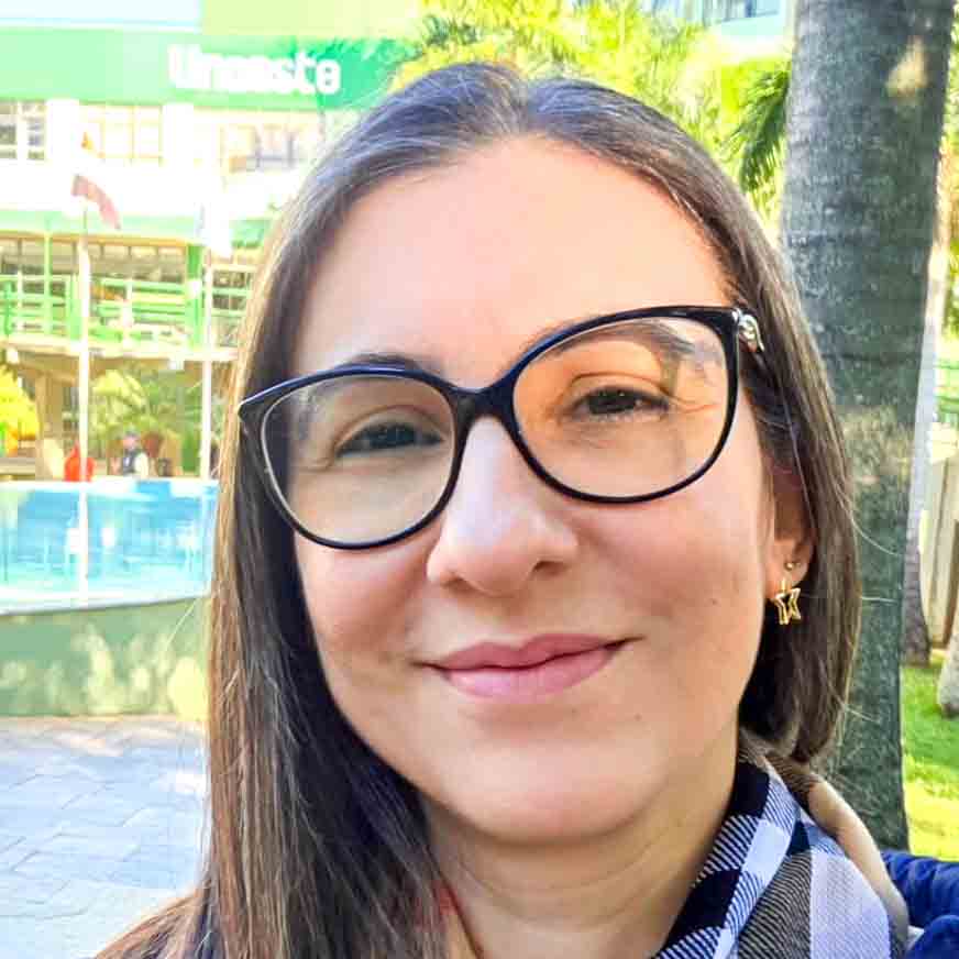 Elisandra, aluna de direito, posando em um ambiente externo com um prédio e uma piscina ao fundo. Ela usa óculos de armação preta e uma echarpe xadrez, com cabelos castanhos e um sorriso amigável.