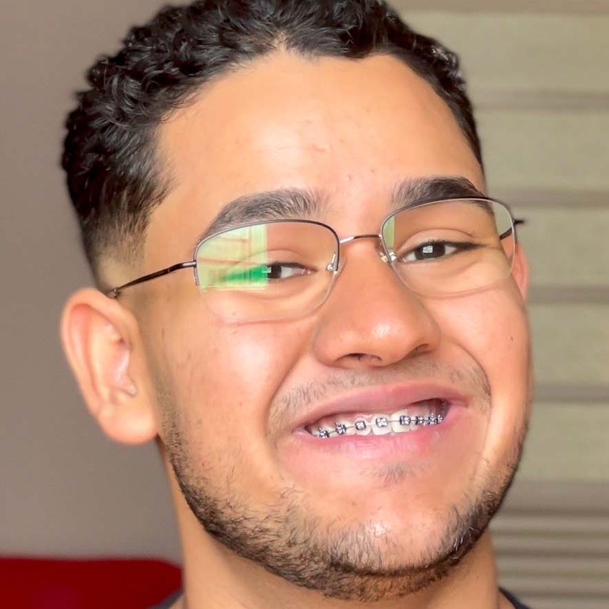 Pedro, aluno de direito, posando em um ambiente interno com um fundo neutro. Ele usa óculos de armação metálica e aparelho ortodôntico, com cabelos curtos e cacheados e um sorriso largo.