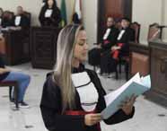 Estudante de Direito da Unoeste participa de júri simulado, usando toga preta com faixa vermelha e segurando documentos azuis. No fundo, colegas caracterizados como membros do tribunal observam atentos. O ambiente remete a um tribunal real, com móveis escuros, microfones e cadeiras estofadas.