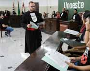 Aluno do curso de Direito da Unoeste realiza júri simulado, trajando toga preta com faixa vermelha e lendo documentos. À sua frente, pessoas sentadas seguram papéis, compondo o júri. Ao fundo, outros estudantes simulam funções judiciais diante de painel verde com o nome da instituição e bandeiras.