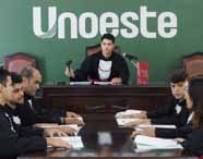 Sala de tribunal simulado na Unoeste, com estudante atuando como juiz ao centro, segurando martelo, rodeado por colegas de toga preta e faixa vermelha. Todos estão sentados à mesa, analisando documentos. O painel verde com "Unoeste" destaca a instituição no cenário acadêmico jurídico.