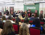 Auditório lotado na Unoeste durante simulação de júri do curso de Direito. Público atento ocupa fileiras de cadeiras vermelhas, enquanto à frente estudantes vestidos de toga participam da atividade. Painel verde com logotipo da Unoeste e bandeiras estaduais compõem o ambiente institucional.