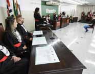 Visão lateral do tribunal simulado na Unoeste, com estudantes de Direito sentados atrás de mesas de madeira, todos de toga preta e faixa vermelha. Uma aluna fala ao microfone, enquanto demais participantes e público acompanham a encenação, evidenciando a estrutura realista do espaço.