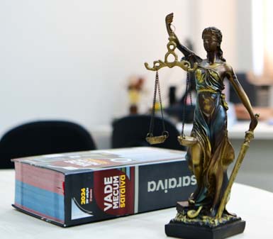Estátua dourada da deusa da Justiça, com balança e espada, posicionada sobre mesa ao lado de um Vade Mecum Saraiva 2024. Ao fundo, ambiente de escritório ou sala de estudos do curso de Direito em Prudente, com cadeiras e objetos desfocados, transmitindo atmosfera acadêmica e jurídica.