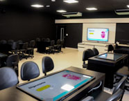 Sala de aula moderna da Unoeste Guarujá, equipada com carteiras pretas, mesas digitais interativas e grande tela ao fundo exibindo conteúdo sobre sistema digestório. O ambiente é tecnológico, com paredes escuras e iluminação clara, voltado para aulas práticas e inovadoras no ensino superior.