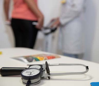 Em consultório de Nutrição, um adipômetro está em destaque sobre a mesa. Ao fundo, uma pessoa de camiseta coral e calça preta realiza avaliação física segurando eletrodos de bioimpedância, enquanto profissional de jaleco branco opera o equipamento. Cena típica do curso de Nutrição no Guarujá.