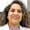 Imagem de Larissa, aluna do curso de fisioterapia no campus Guarujá, trajando jaleco branco. O fundo apresenta elementos institucionais com cores claras, reforçando o contexto acadêmico e profissional.