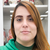 Imagem de Maria, aluna do curso de fisioterapia no campus Guarujá, vestindo uma blusa verde. O fundo apresenta prateleiras brancas, remetendo a um ambiente institucional moderno e organizado.