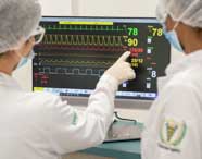 Dois estudantes de Fisioterapia analisam monitor multiparamétrico em laboratório, vestindo jalecos brancos, toucas e máscaras. Um deles aponta para os gráficos coloridos do monitor, interpretando dados de frequência cardíaca, pressão arterial e saturação de oxigênio
