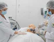 Três estudantes de Fisioterapia da Unoeste, com jalecos brancos, toucas e máscaras, realizam atendimento simulado em manequim adulto. Uma aluna executa ventilação manual enquanto as outras acompanham o procedimento, demonstrando trabalho em equipe no laboratório.