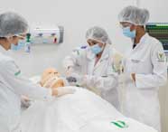 Grupo de três estudantes de Fisioterapia, com jalecos e toucas, participa de simulação de suporte respiratório em manequim adulto. Uma aluna opera o ambu, outra auxilia e a terceira observa, enquanto monitor cardíaco exibe sinais vitais ao fundo.