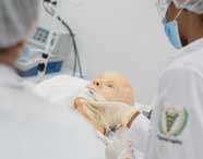 Cena mostra duas estudantes de Fisioterapia em simulação clínica, ambas com jalecos brancos e máscaras, realizando procedimentos de ventilação em manequim adulto deitado, reforçando práticas de emergência respiratória em laboratório.