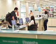 Biblioteca universitária ampla e iluminada, com estantes repletas de livros acadêmicos ao fundo. Alunos utilizam computadores no balcão de atendimento, outros circulam pelo espaço, evidenciando ambiente de estudo e pesquisa da instituição.