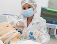 Aluna de Fisioterapia realiza procedimento de intubação orotraqueal em manequim adulto, utilizando ambu azul para ventilação. Ela veste jaleco branco, luvas e touca, enquanto monitor cardíaco exibe sinais vitais ao fundo, em laboratório de simulação realística.