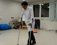 Aluno de Fisioterapia em jaleco branco caminhando com muletas em um laboratório ou clínica, demonstrando reabilitação, com equipamentos de fisioterapia ao redor.