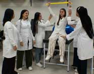 Grupo de alunas de Fisioterapia em jalecos brancos realizando uma aula prática interativa com bastões e exercitadores em um ambiente de laboratório.