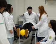 Alunos de Fisioterapia em jalecos brancos realizando uma aula prática de reabilitação, com um aluno deitado em uma maca e outros observando e auxiliando.