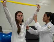 Três alunas de Fisioterapia em jalecos brancos realizando um exercício de alongamento com faixa elástica amarela, com uma professora orientando os movimentos.
