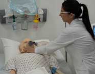 Professora ou aluna de Fisioterapia em jaleco branco utilizando um ventilador mecânico em um manequim de paciente para simulação de tratamento respiratório.