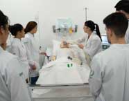 Grupo de alunos de Fisioterapia em jalecos brancos observando e aprendendo sobre ventilação mecânica com um manequim de paciente e equipamentos médicos.