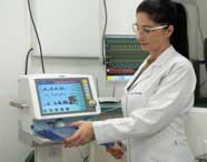 Professora de Fisioterapia em jaleco branco com óculos, operando um ventilador mecânico e registrando dados em uma prancheta em ambiente de simulação.