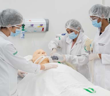 Três estudantes do curso de Fisioterapia da Uniesp Guarujá, usando jalecos brancos, toucas e luvas, realizam treinamento prático em laboratório com manequim hospitalar adulto deitado em maca. Eles simulam procedimentos de atendimento respiratório em ambiente clínico equipado.