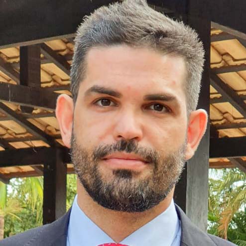 Gabriel, aluno de Educação Física - Bacharelado, posando em um ambiente externo com uma estrutura de madeira ao fundo. Ele tem barba e cabelos grisalhos, e usa um terno, com uma expressão séria.