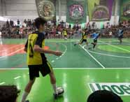Partida de futsal durante festival esportivo no ginásio da Unoeste Guarujá. Atletas com uniformes amarelos e pretos enfrentam equipe de azul claro e preto. A quadra possui piso verde e laranja, com público atento nas arquibancadas e banners coloridos decorando as paredes.