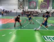 Jogo de futsal entre equipes universitárias na Unoeste Guarujá. Estudantes com uniformes azul e amarelo enfrentam time de preto e laranja, disputando a bola em quadra verde e laranja. Arquibancada cheia e faixas temáticas ao fundo reforçam o clima de competição acadêmica.