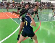 Partida de handebol feminino em andamento na quadra da Unoeste Guarujá. Aluna de camisa azul prepara arremesso enquanto atleta adversária de uniforme preto e verde tenta bloquear. Outras jogadoras acompanham a jogada e o público observa atentamente das arquibancadas.