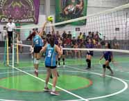 Equipe feminina de vôlei disputa ponto em quadra da Unoeste Guarujá. Jogadora de uniforme azul salta para atacar a bola, enquanto colegas e adversárias de roxo acompanham a jogada. A rede divide os times e o público assiste das arquibancadas ao fundo, em ambiente animado.