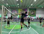 Partida masculina de vôlei no festival esportivo da Unoeste Guarujá. Atleta de uniforme preto e verde executa um ataque potente, saltando próximo à rede, enquanto adversários tentam bloquear. Banners coloridos e público nas arquibancadas compõem o cenário dinâmico do evento.