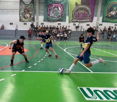 Jogo de futsal em quadra coberta, com estudantes de Educação Física da Unoeste Guarujá. Atletas usam uniformes azul e amarelo ou preto e laranja, disputando a bola em movimento intenso. Ao fundo, arquibancada cheia de espectadores e bandeiras coloridas decoram as paredes do ginásio.