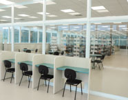 Sala de estudos da biblioteca do curso de Psicologia da Unoeste Guarujá, com cabines individuais de madeira clara e cadeiras pretas. Ambiente envidraçado, permitindo visão para o interior da biblioteca, onde há mesas redondas, cadeiras e estantes cheias de livros. Espaço claro, moderno e silencioso, ideal para concentração dos estudantes.