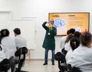 Professor de Psicologia ou Neurociência em jaleco verde demonstrando um modelo anatômico do cérebro para alunos em sala de aula, com material didático digital exibido em tela.