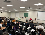 Auditório da Unoeste Guarujá durante evento acadêmico do curso de Psicologia. Estudantes ocupam fileiras de cadeiras, atentos à mesa composta por palestrantes e professores. Ao fundo, quadro verde, projetor e painel com o nome da instituição. Ambiente amplo, iluminado, propício para debates, palestras e atividades universitárias.