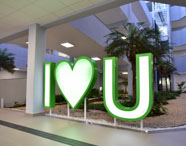 Escultura luminosa com letras “I”, coração e “U” em verde e branco, localizada no jardim interno do campus da Unoeste Guarujá. Ao fundo, palmeiras e canteiros de plantas, sob marquise moderna e iluminação clara. Espaço transmite acolhimento, integração e identidade visual marcante da instituição de ensino superior.