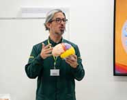 Professor de Psicologia com jaleco verde e óculos apresentando e explicando as diferentes partes de um modelo anatômico colorido do cérebro em sala de aula.