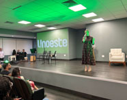 Auditório da Unoeste Guarujá durante apresentação, com professora em destaque no palco, segurando microfone. Público atento nas poltronas, ambiente moderno com iluminação verde, telão exibindo logotipo da instituição. No palco, instrumentos musicais e decoração discreta. Espaço propício para eventos, palestras e apresentações acadêmicas.