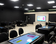 Sala de aula tecnológica do campus Unoeste Guarujá, equipada com mesas digitais interativas, cadeiras pretas e lousa digital ao fundo. Ambiente moderno, paredes escuras, iluminação branca e ar-condicionado. Estrutura voltada para atividades práticas e dinâmicas, atendendo às necessidades dos cursos de saúde e tecnologia.