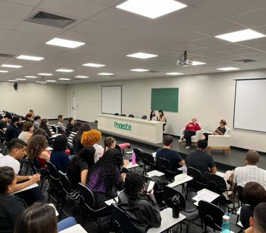 Auditório universitário da Unoeste com estudantes do curso de Psicologia assistindo a uma palestra. No palco, duas pessoas sentadas em poltronas brancas participam de um bate-papo, enquanto outras três estão atrás de uma bancada com o nome da instituição. Ambiente moderno, com iluminação clara e quadro verde ao fundo.
