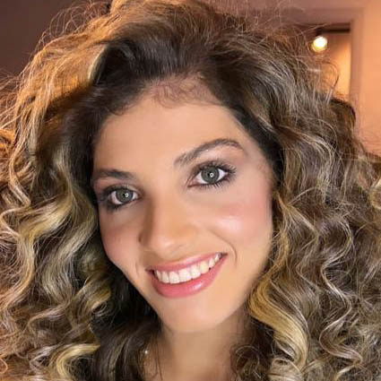 Ana, aluna de Nutrição, posando em um ambiente interno com iluminação quente ao fundo. Ela tem cabelos cacheados e olhos verdes, com um sorriso largo.