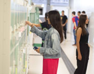 Estudantes da Unoeste Jaú utilizam armários individuais em corredor claro e moderno. Uma aluna de jaqueta jeans e calça rosa mexe no armário enquanto outros alunos conversam ao fundo. O ambiente é organizado, com sinalização tátil no piso e atmosfera dinâmica.