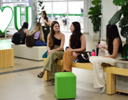 Estudantes sentados em bancos de madeira claros e cubos verdes nas áreas de convivência da Unoeste Jaú. Ao fundo, letreiro luminoso “I ♥ U”, plantas decorativas e portas de vidro, compondo um espaço confortável e vibrante para interação entre alunos.