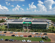 Foto aérea do campus Unoeste Jaú, edifício moderno com fachada de vidro espelhado, painéis solares no telhado e nome Unoeste em destaque. Amplo estacionamento com carros alinhados, área verde ao redor e campos agrícolas ao fundo, mostrando a integração com a paisagem local.