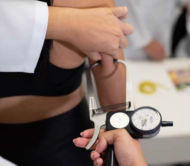Close de avaliação antropométrica realizada por estudante ou profissional do curso de Nutrição, utilizando adipômetro para medir a dobra cutânea no braço de uma pessoa. O cenário é uma sala clara, com jaleco branco e instrumentos típicos de práticas acadêmicas em instituições de ensino superior.