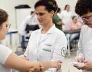 Em sala de aula prática do curso de Fisioterapia da Unoeste, estudantes com jaleco branco realizam atendimento clínico simulado. Uma aluna segura o braço de uma paciente enquanto outro estudante opera um aparelho eletrônico. Ao fundo, outros alunos acompanham a atividade, todos em ambiente iluminado e organizado.