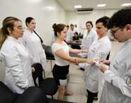 Grupo de estudantes do curso de Fisioterapia da Unoeste, usando jalecos brancos, participam de aula prática em clínica. Uma paciente está de pé, com eletrodos nas pernas e quadril, sendo avaliada por colegas que seguram seus braços e utilizam equipamentos eletrônicos. Ambiente limpo, com macas e ar-condicionado visíveis ao fundo.