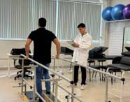 Na clínica de fisioterapia da Unoeste, estudante com jaleco branco anota informações em prancheta enquanto paciente realiza caminhada entre barras paralelas. O ambiente possui bolas de pilates coloridas, macas e iluminação natural vinda das janelas, evidenciando a estrutura moderna do local.
